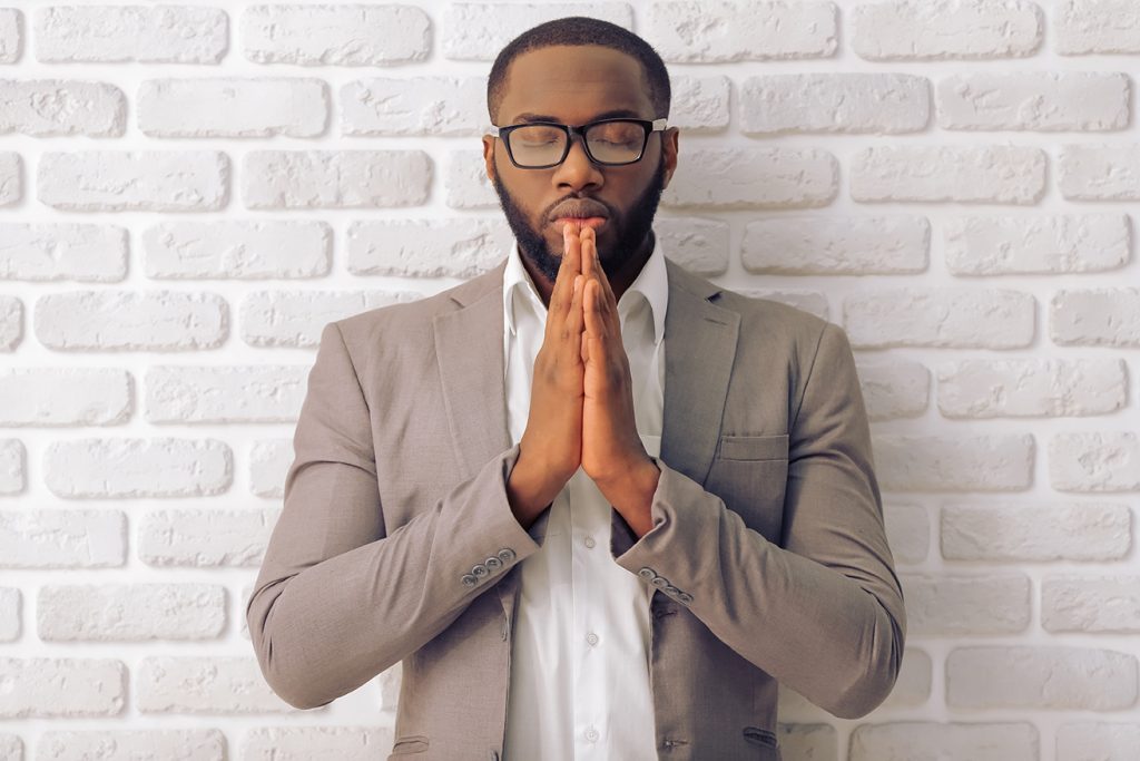 Black man in suit praying against a white brick wall